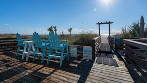 A wooden deck with blue chairs and a table, overlooking a beach with palm trees and a wooden pier. - Top of the Gulf 320 Beachfront Free Beach Chairs (Panama City Beach)