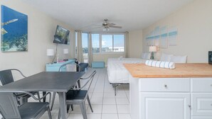 A hotel room with a bed, a desk with a chair, a TV, a ceiling fan, and a window with a view of the city. - Top of the Gulf 320 Beachfront Free Beach Chairs (Panama City Beach)