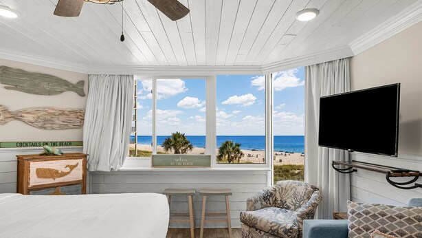 A bedroom with a large bed, a TV mounted on the wall, a window with a view of the beach, and a ceiling fan. - Beachfront Studio Condo - Top of the Gulf 317 (Panama City Beach)