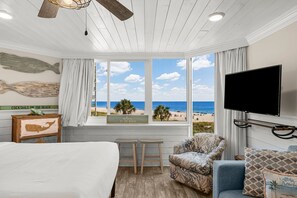 Une chambre avec une grande fenêtre donnant sur la plage, un lit, une chaise et un téléviseur.