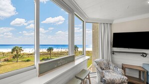 A room with a large window overlooking a beach, a flat-screen TV mounted on the wall, and a patterned sofa. - Beachfront Studio Condo - Top of the Gulf 317 (Panama City Beach)