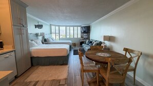 A compact room with a bed, a small sofa, a wooden dining table with chairs, a kitchenette, and a large window providing natural light. - Renovated Beach Front Condo - Top of the Gulf 302 (Panama City Beach)