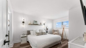 A bedroom with a large bed, bedside tables, a nightstand, a chair, a lamp, a shelf with decorative items, and a window with blinds. - Summit 808 - Luxury Beach Resort Condo 1-6 People (Panama City Beach)