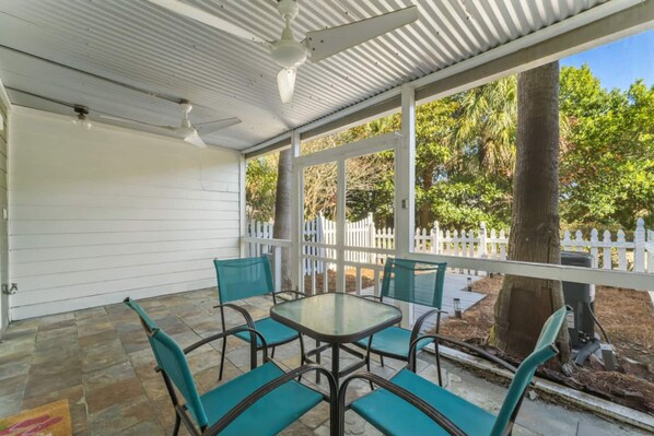 A covered patio with a table and chairs, a white fence, and trees in the background.
