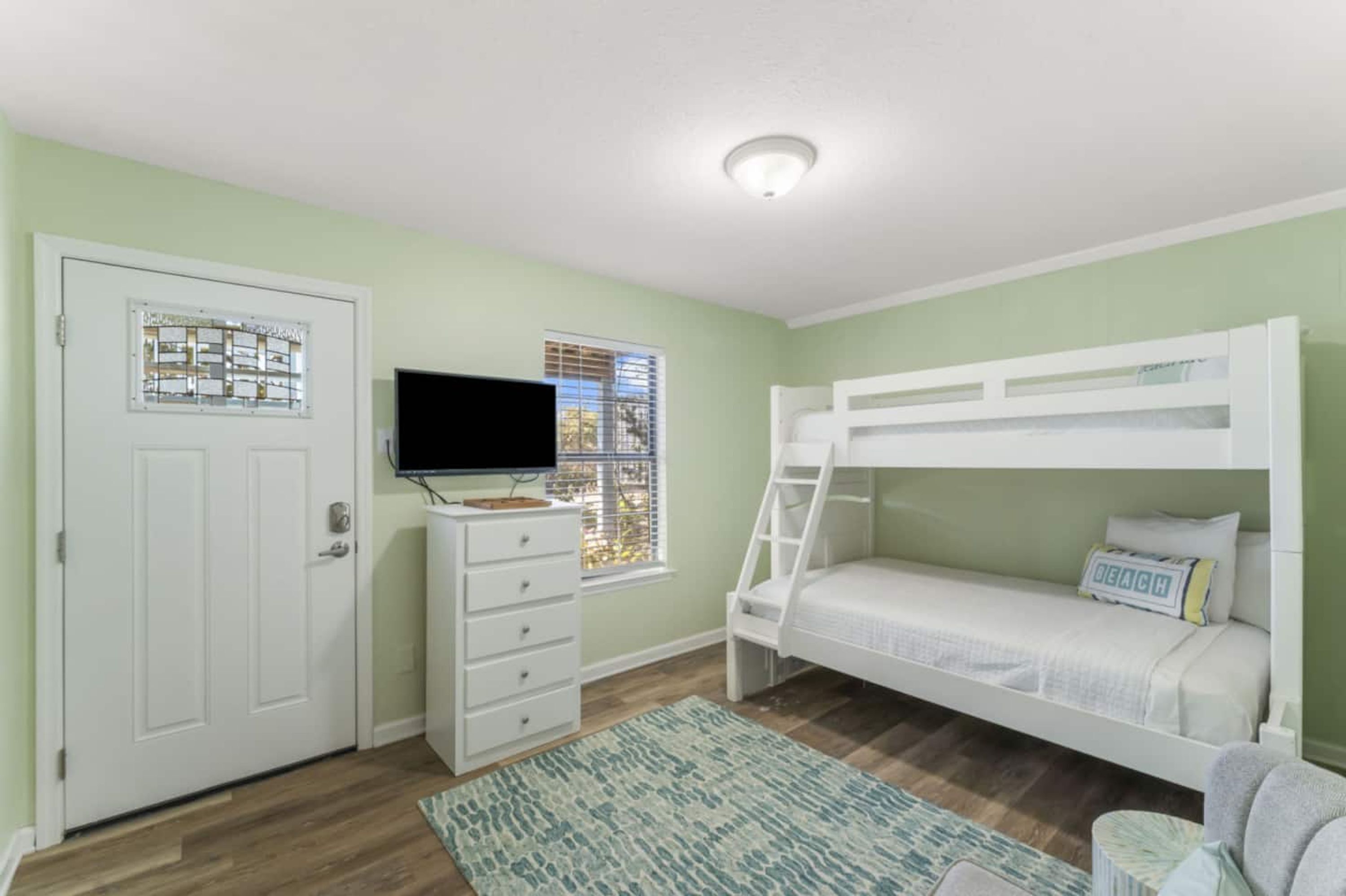 Une chambre avec un lit superposé, une commode, un téléviseur et une fenêtre avec des stores.