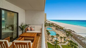 A balcony with a wooden table and chairs, a sofa, and a large window overlooking a beach with a pool area.