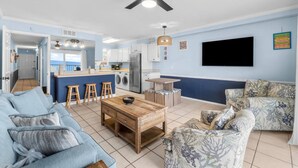 A modern living room with a sofa, a coffee table, and a patterned armchair. A kitchen area with a refrigerator, sink, and bar stools is visible in the background. - Summit 505 Beach Resort Condo - Balcony Best View (Panama City Beach)