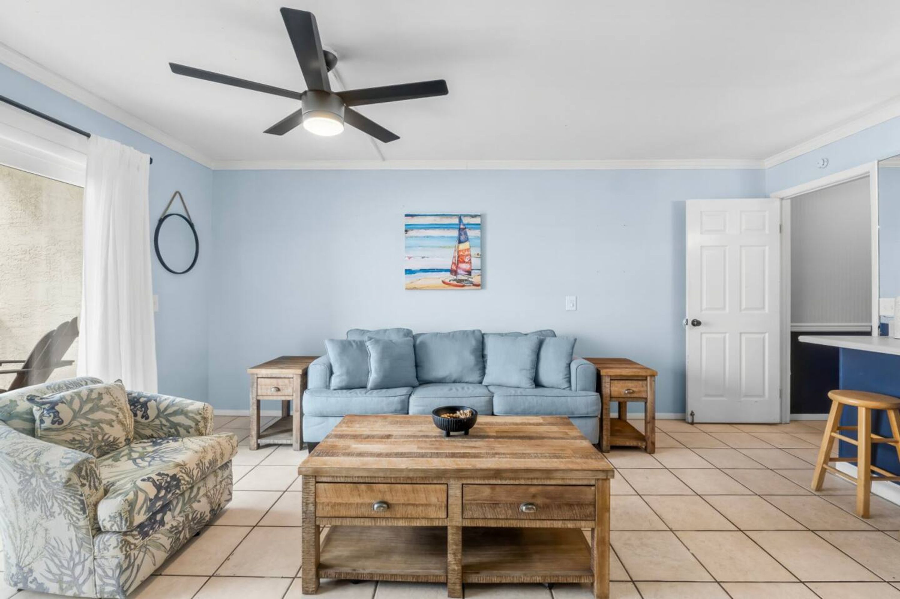 Ein Wohnzimmer mit einem blauen Sofa, einem Holztisch, einem gemusterten Sessel, einem Deckenventilator und einem Bild an der Wand.
