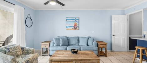 Ein Wohnzimmer mit einem blauen Sofa, einem Holztisch, einem gemusterten Sessel, einem Deckenventilator und einem Bild an der Wand.