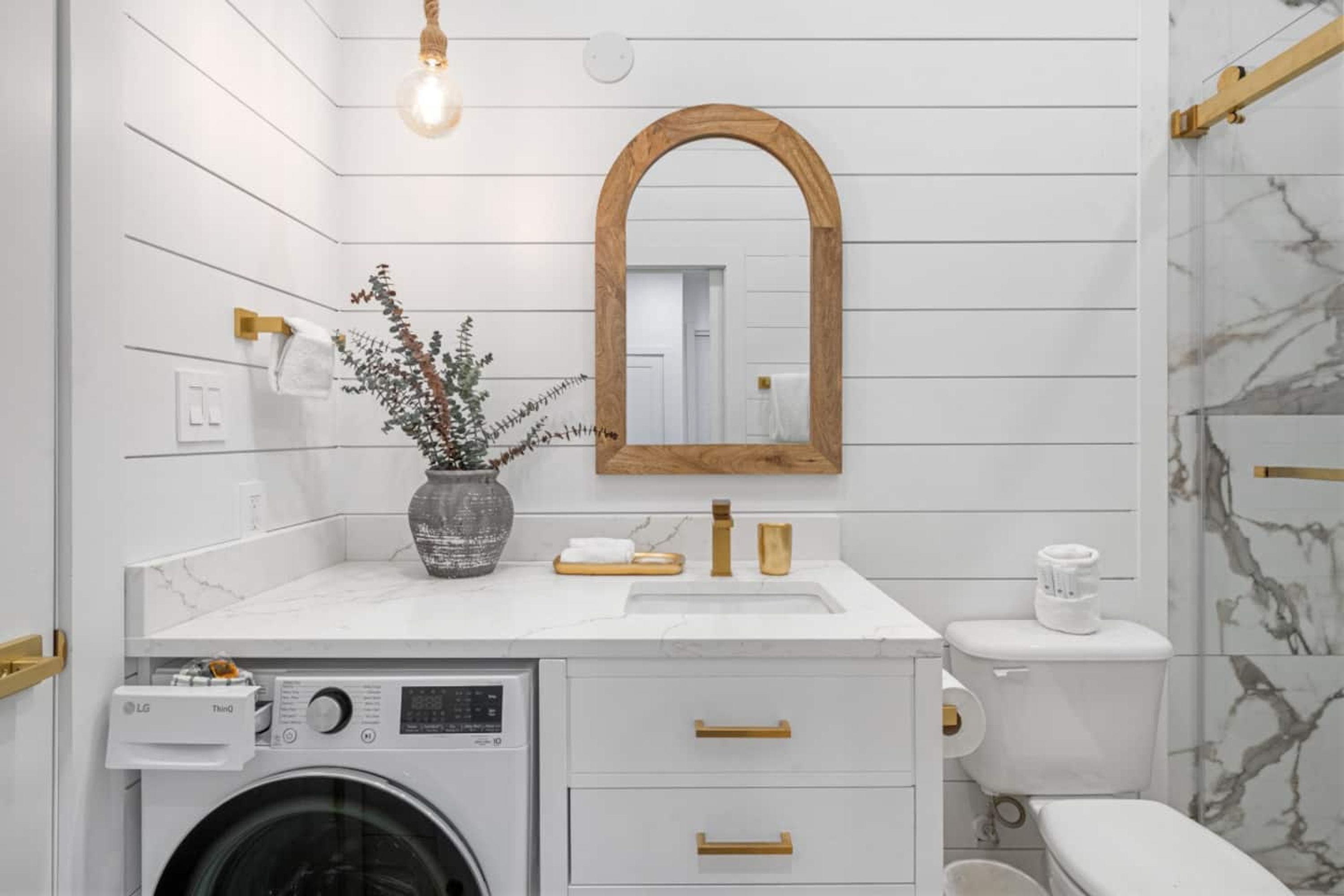 A modern bathroom with a washing machine, a sink with gold handles, a wooden-framed mirror, and marble-patterned walls.