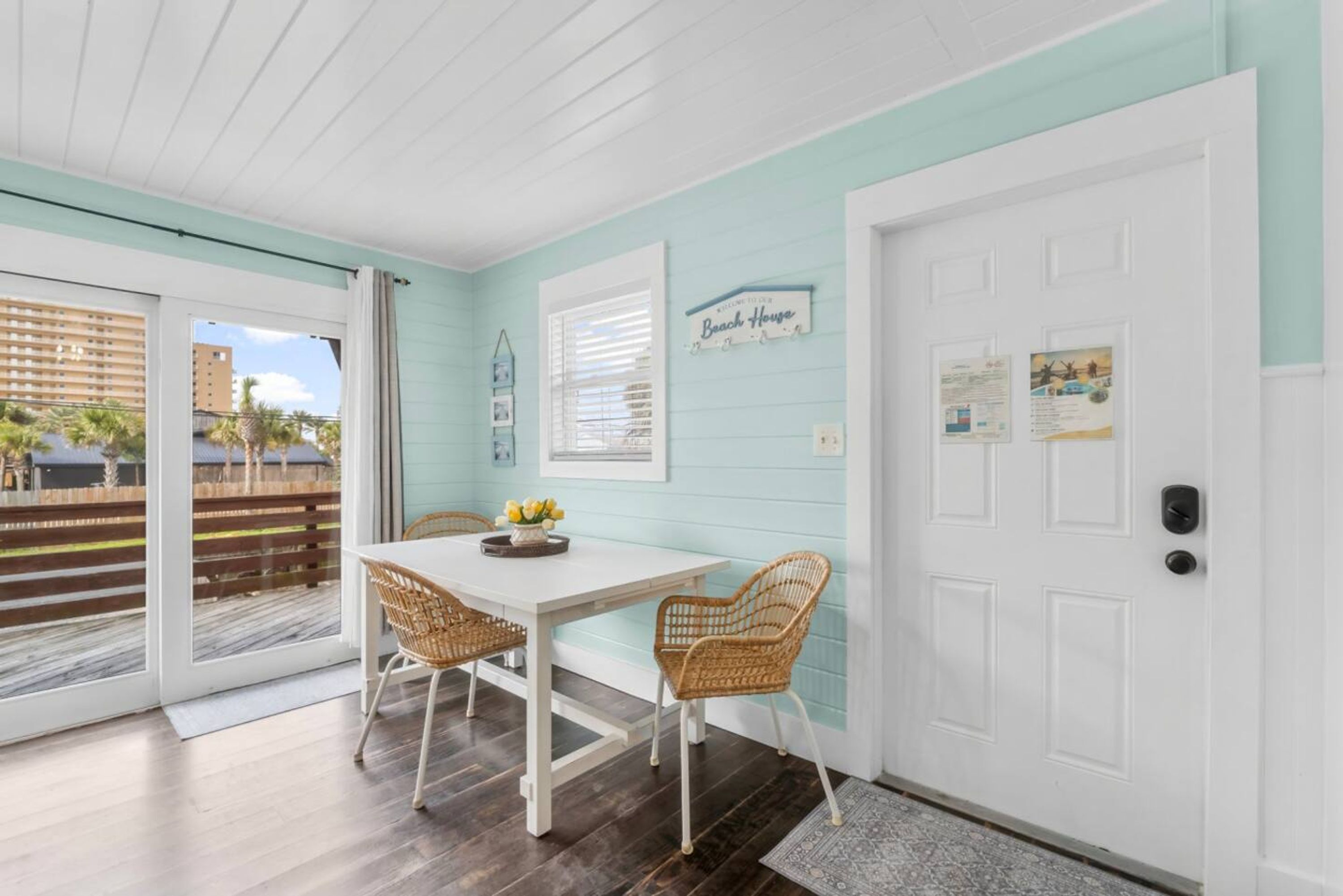 Et værelse med et hvidt bord og fletstole, en hvid dør med lås, og et skilt med teksten "Welcome to the Beach House".