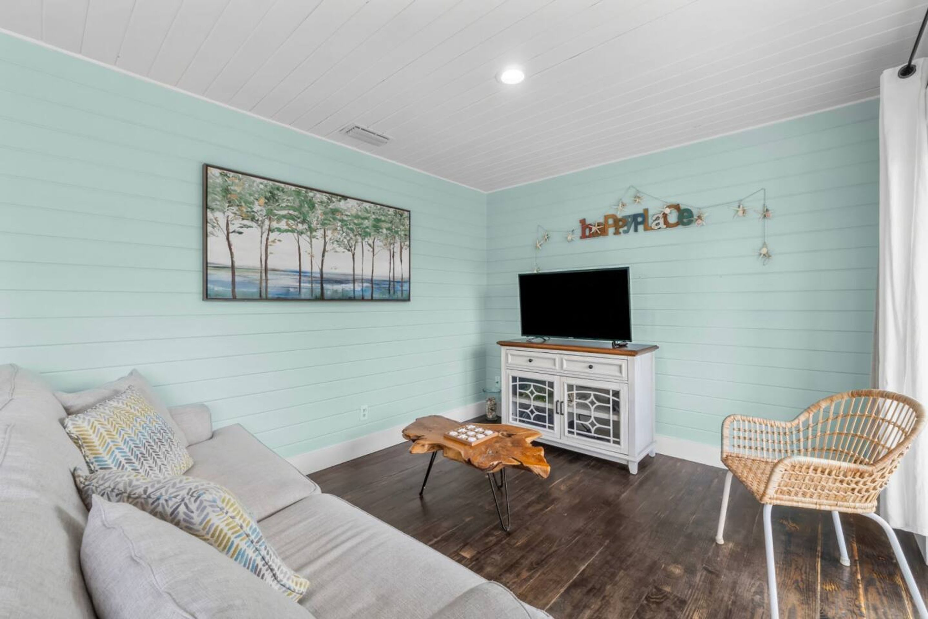 A living room with a light blue wall, a wooden coffee table, a grey sofa, a wicker chair, and a TV on a white cabinet.