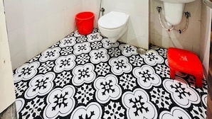 Un baño con inodoro, lavabo y ducha. Hay un cubo y un taburete, ambos rojos. El piso tiene un estampado floral en blanco y negro.
