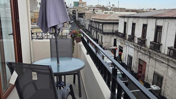 Un balcón con una mesa redonda y sillas, una sombrilla morada, y vistas de la ciudad.
