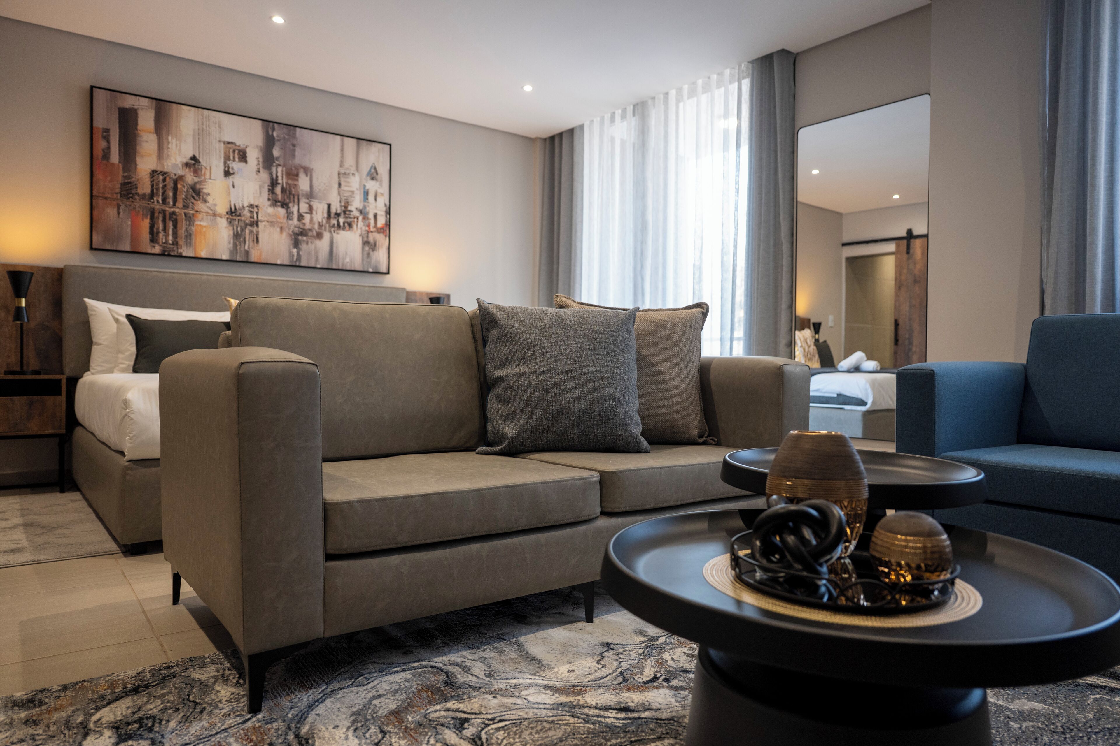 A modern hotel room with a sectional sofa, a round coffee table with decorative items, a large abstract painting, and a large mirror.