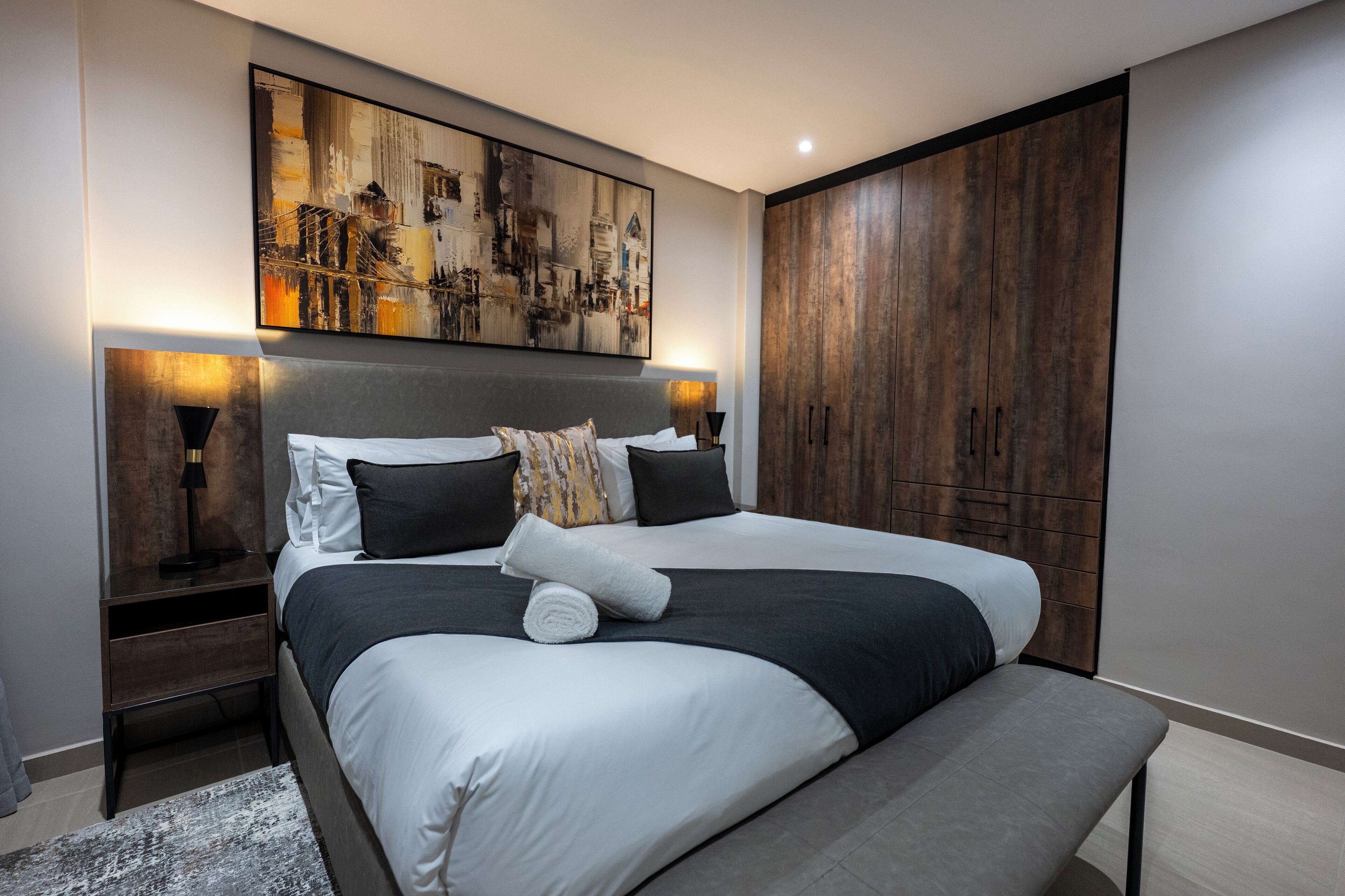 A modern bedroom with a large bed, a headboard with artwork, bedside lamps, and a wooden wardrobe.