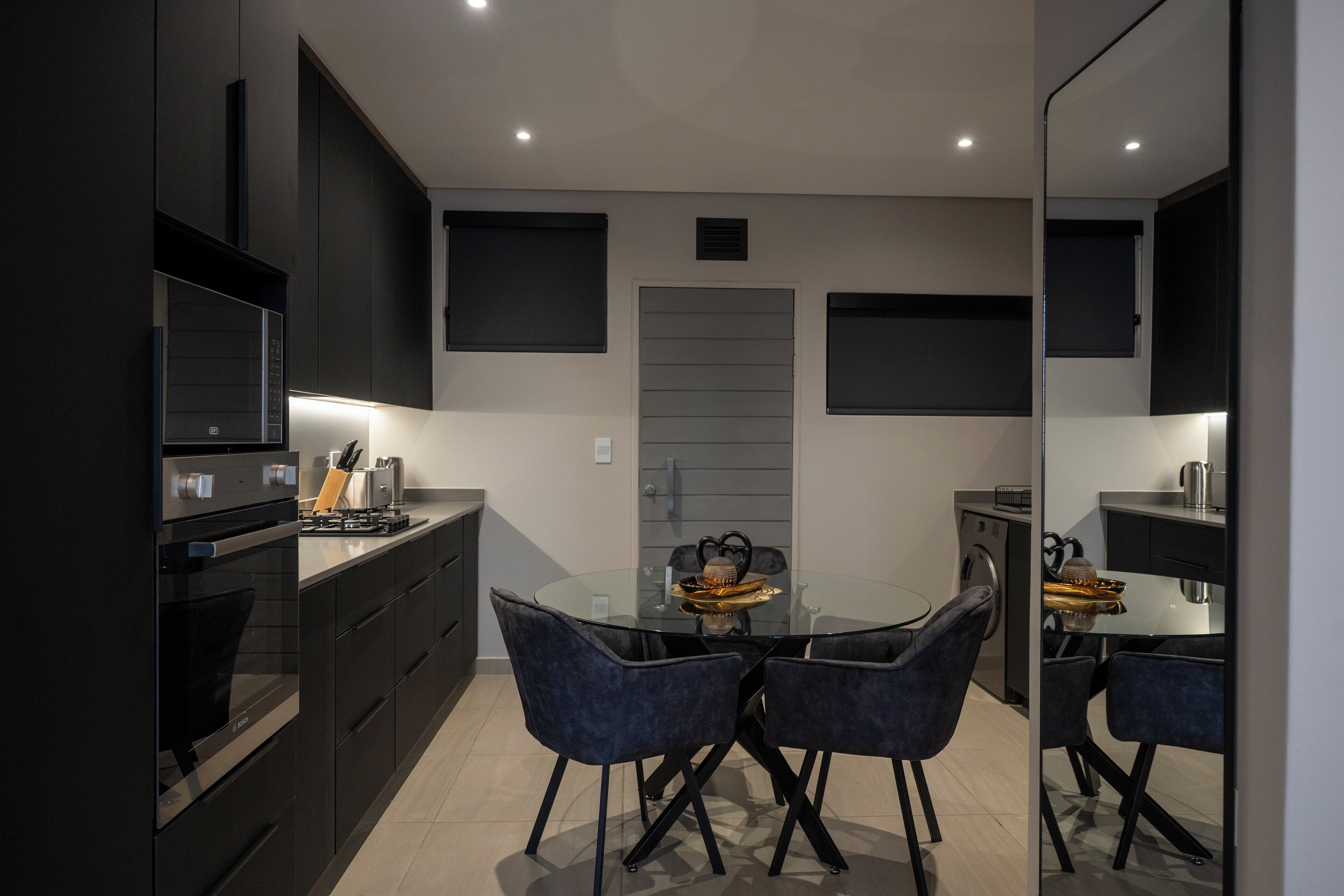 A modern kitchen with dark cabinetry, a glass dining table, and black chairs.
