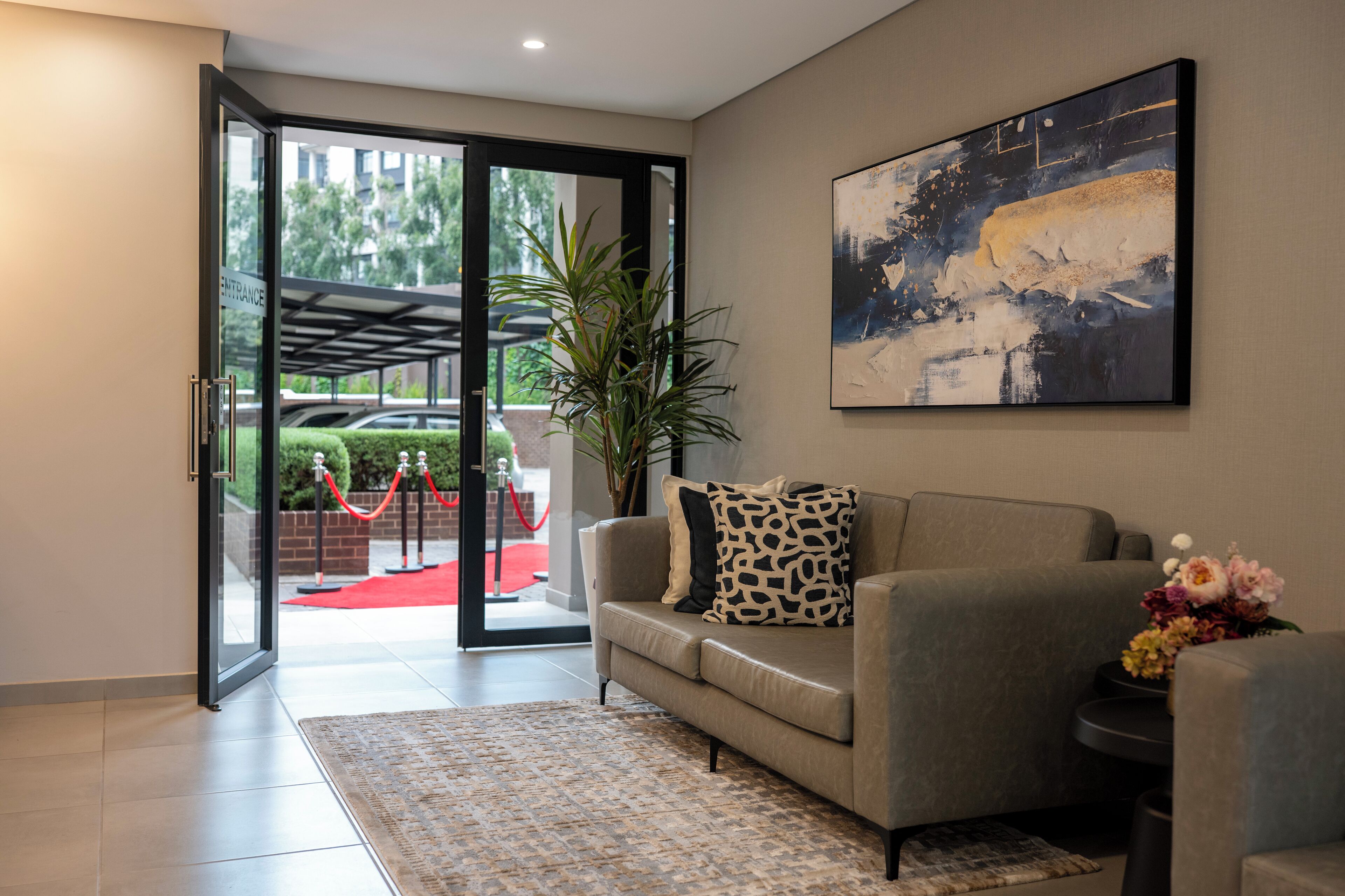A modern hotel lobby with a sofa, a large abstract painting, a potted plant, and a red carpet leading outside.