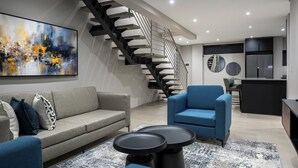 A modern living room with a grey sofa, blue armchair, and a black coffee table. A staircase with a black railing leads to a kitchen area with a refrigerator and a circular mirror. - Lux Leisure Apartment Hotel Sandton (Sandton)