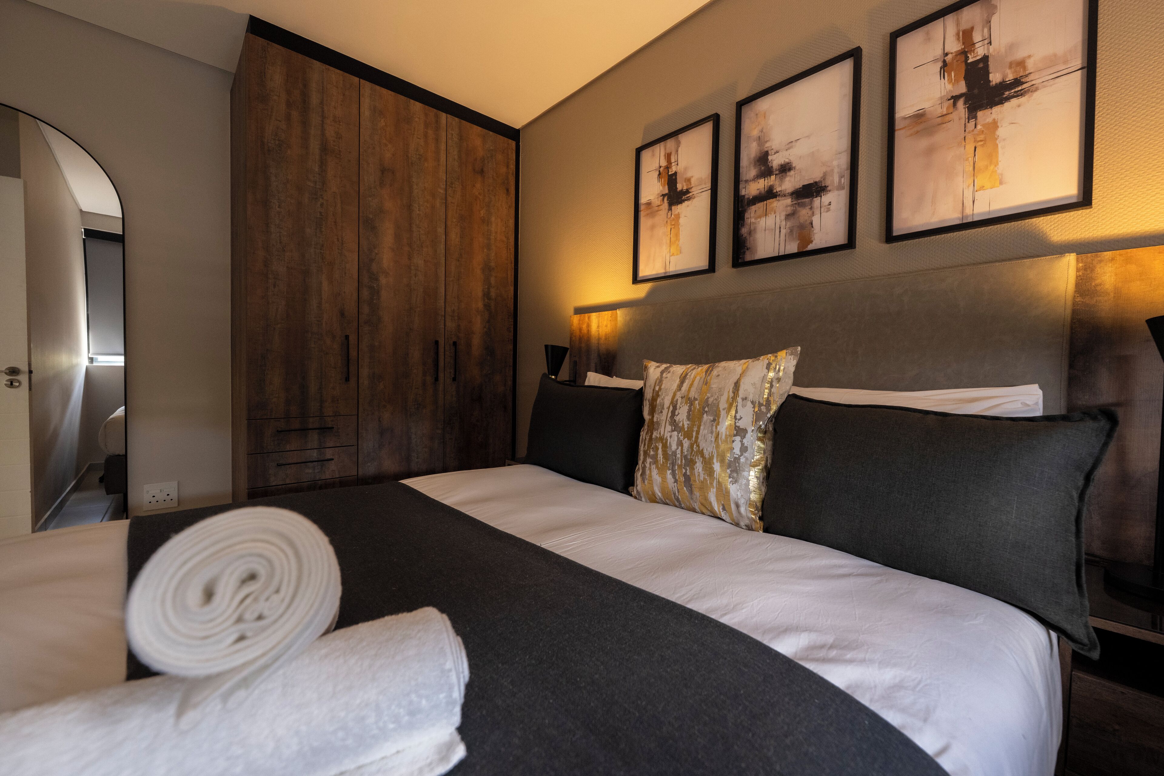 A neatly made bed with a dark headboard, a large wooden wardrobe, and abstract art on the wall.