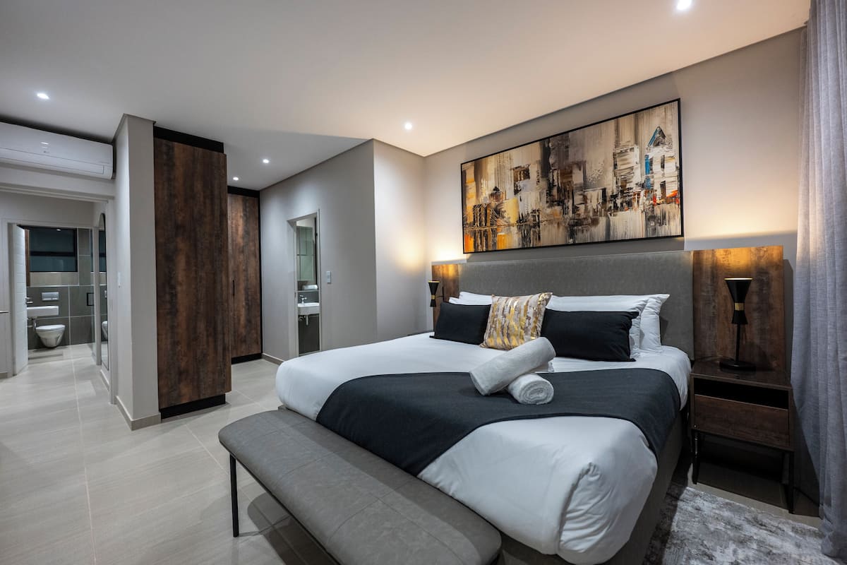 Un dormitorio moderno con una cama grande, un banco, un cuadro en la pared y un baño a la vista al fondo.