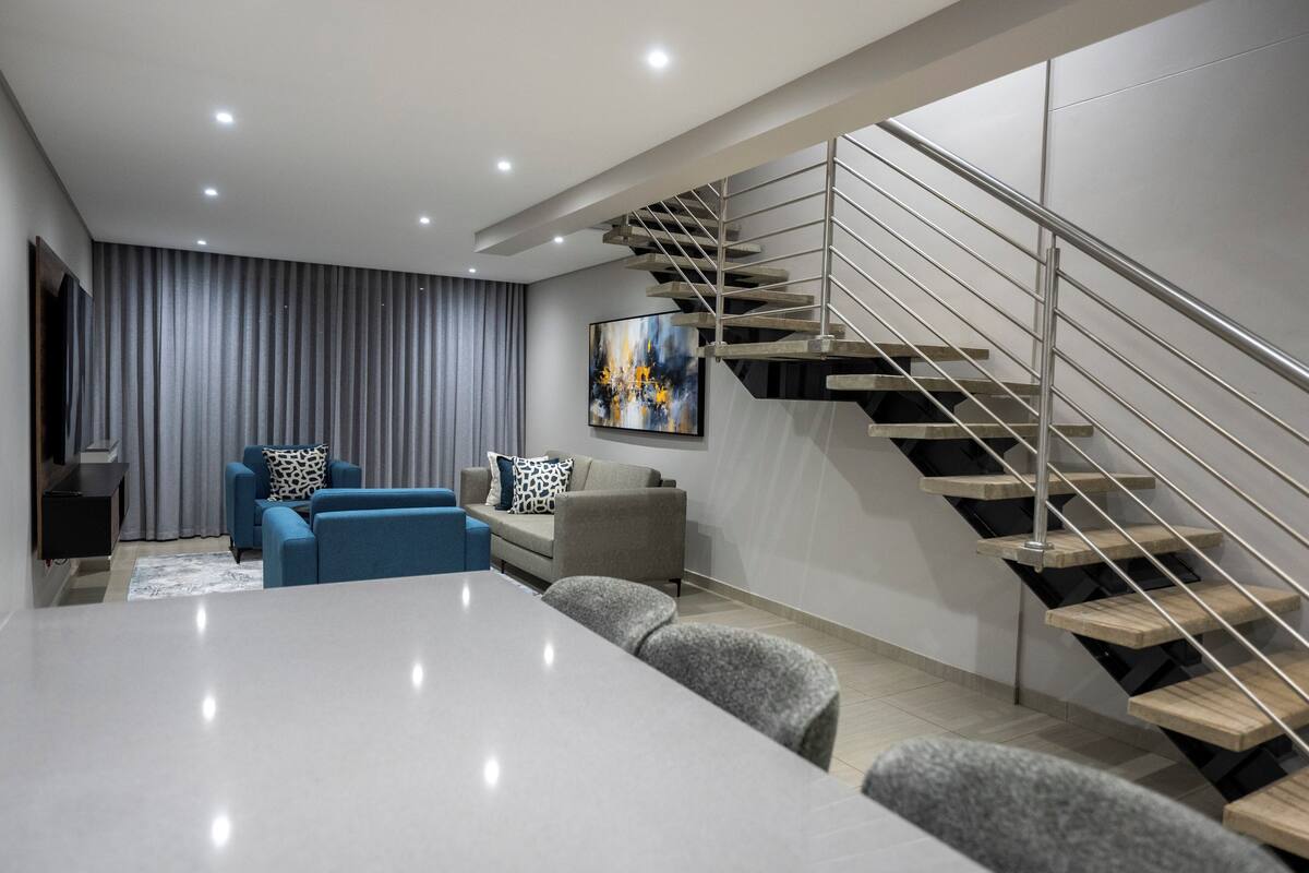Una sala de estar moderna con una mesa blanca grande, sofás azules, un sofá gris y una escalera con barandilla de metal.