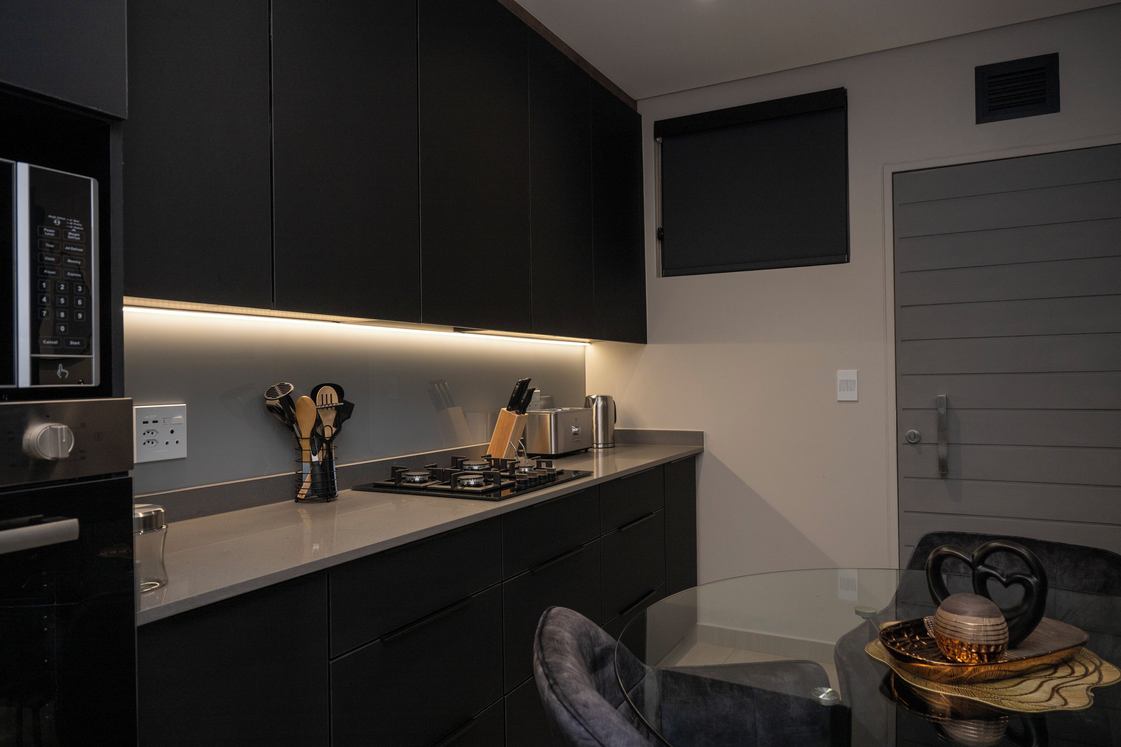 Une cuisine moderne avec des meubles de couleur sombre, un four encastré et une cuisinière à gaz. On y trouve une table à manger en verre avec une chaise, ainsi qu’un petit objet décoratif posé sur la table.
