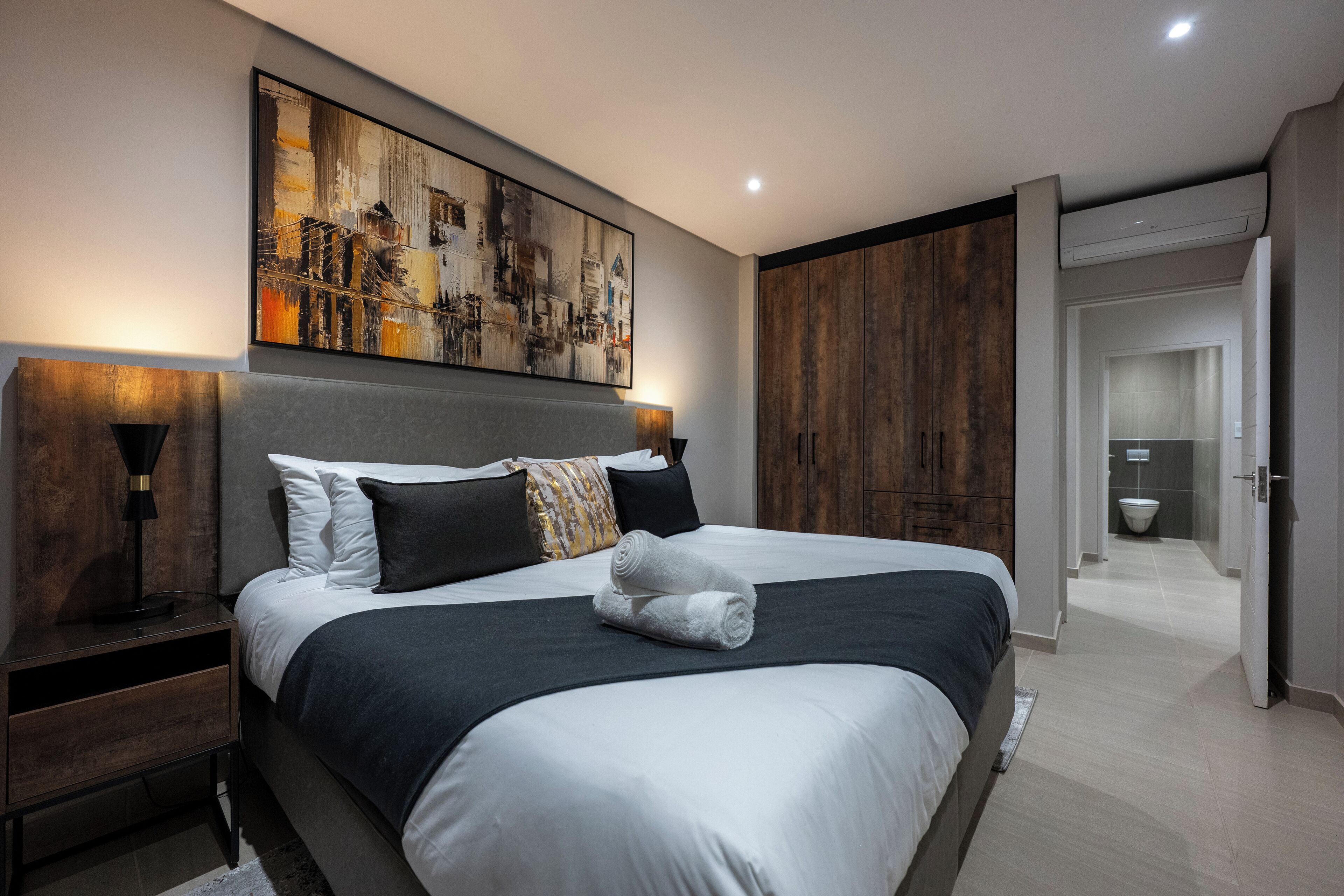 A modern bedroom with a large bed, a headboard with a cityscape painting, a nightstand with a lamp, and a wooden wardrobe.