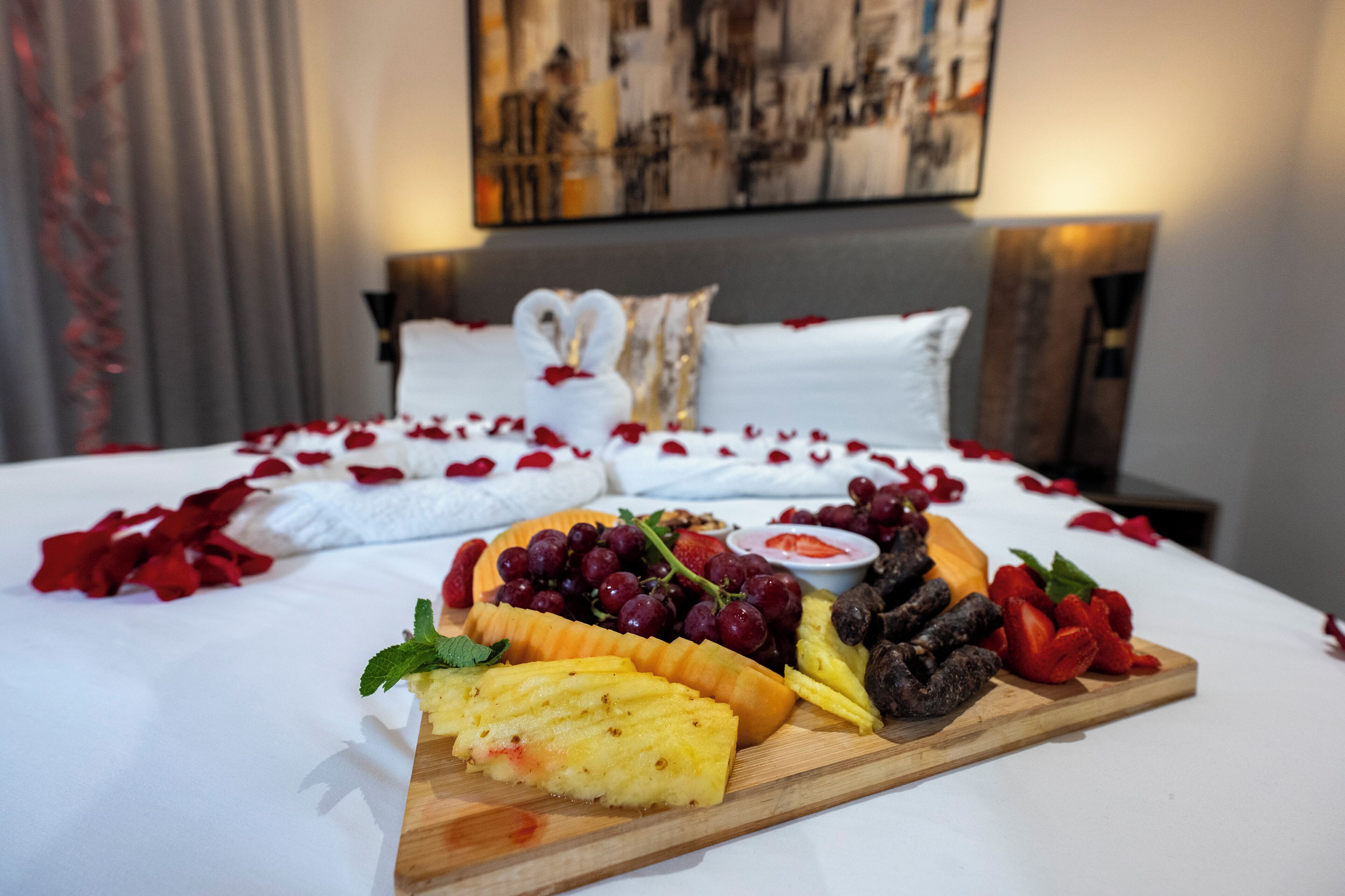 Una cama con sábanas blancas y pétalos de rosa, una tabla de madera con frutas y queso, y un cuadro en la pared.