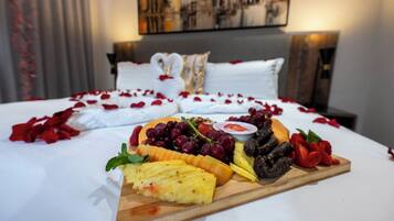 Una cama con sábanas blancas y pétalos de rosa, una tabla de madera con frutas y queso, y un cuadro en la pared.