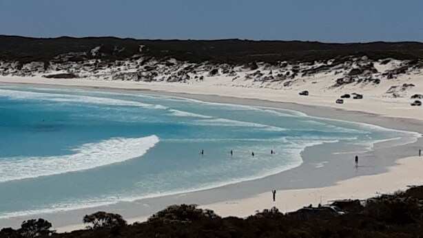 En strand med klart blåt vand, sandet bredde og klippeformationer.