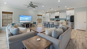 A modern living room with a sofa, coffee table, and TV. It includes a kitchen with bar stools and a refrigerator. - Introducing The Barefoot Villa in Lost Key! For An Upscale-Coastal Vacation. (Pensacola)