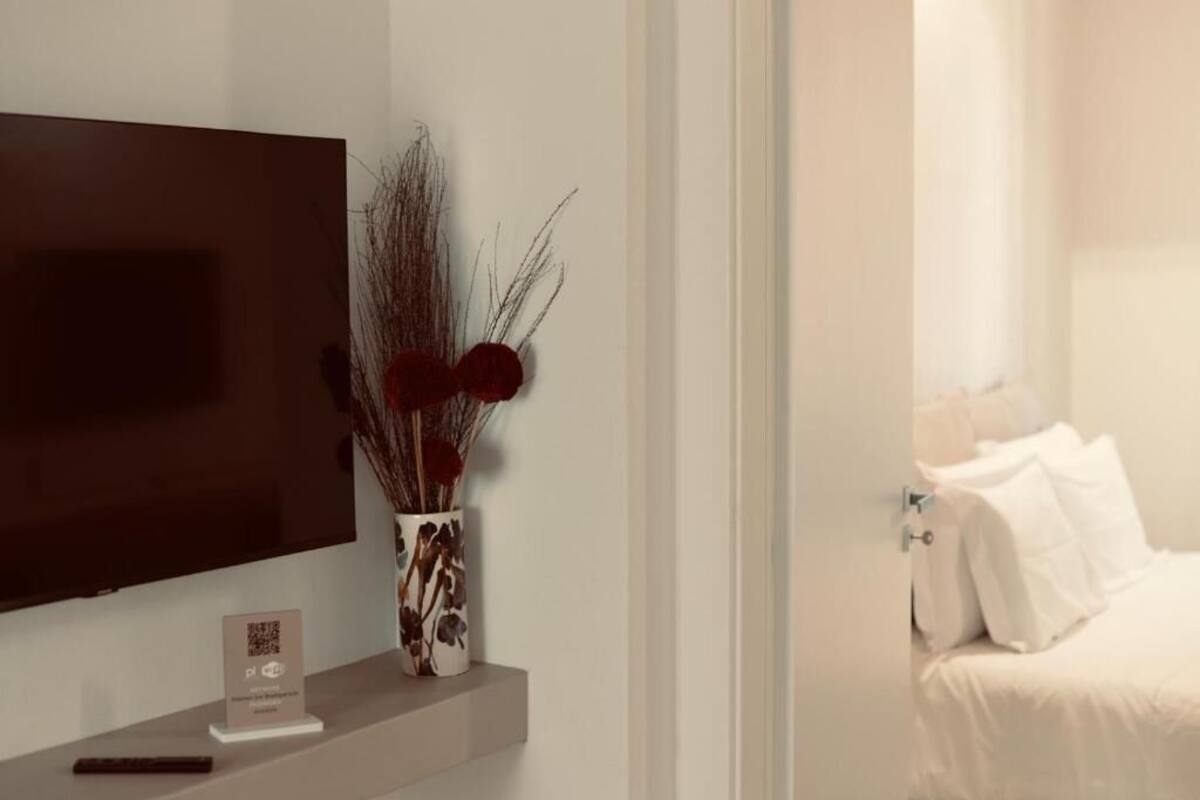 Una habitación de hotel moderna con una televisión de pantalla plana grande montada en la pared, una mesita de noche con un jarrón de plantas secas, y una cama bien hecha con ropa blanca.