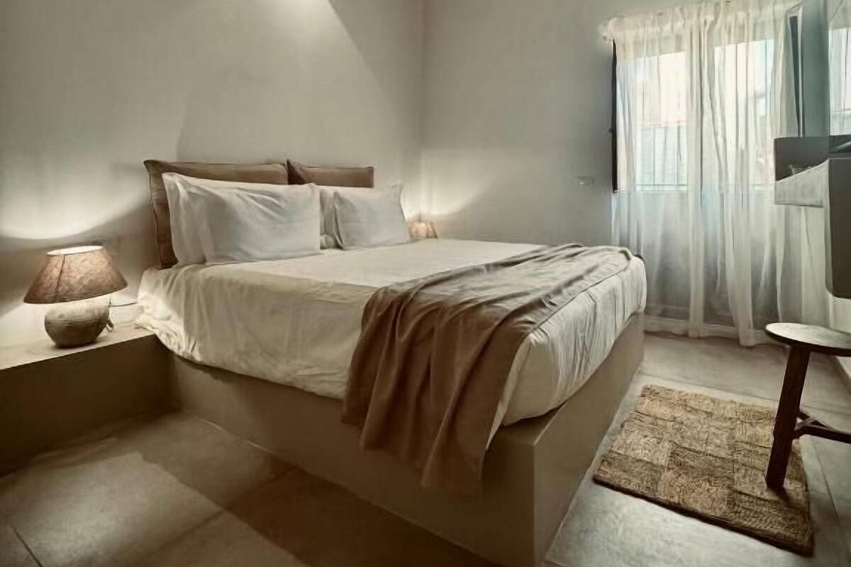 Un dormitorio con techo de madera, una cama con ropa blanca, una lámpara de noche y una mesita con una silla.