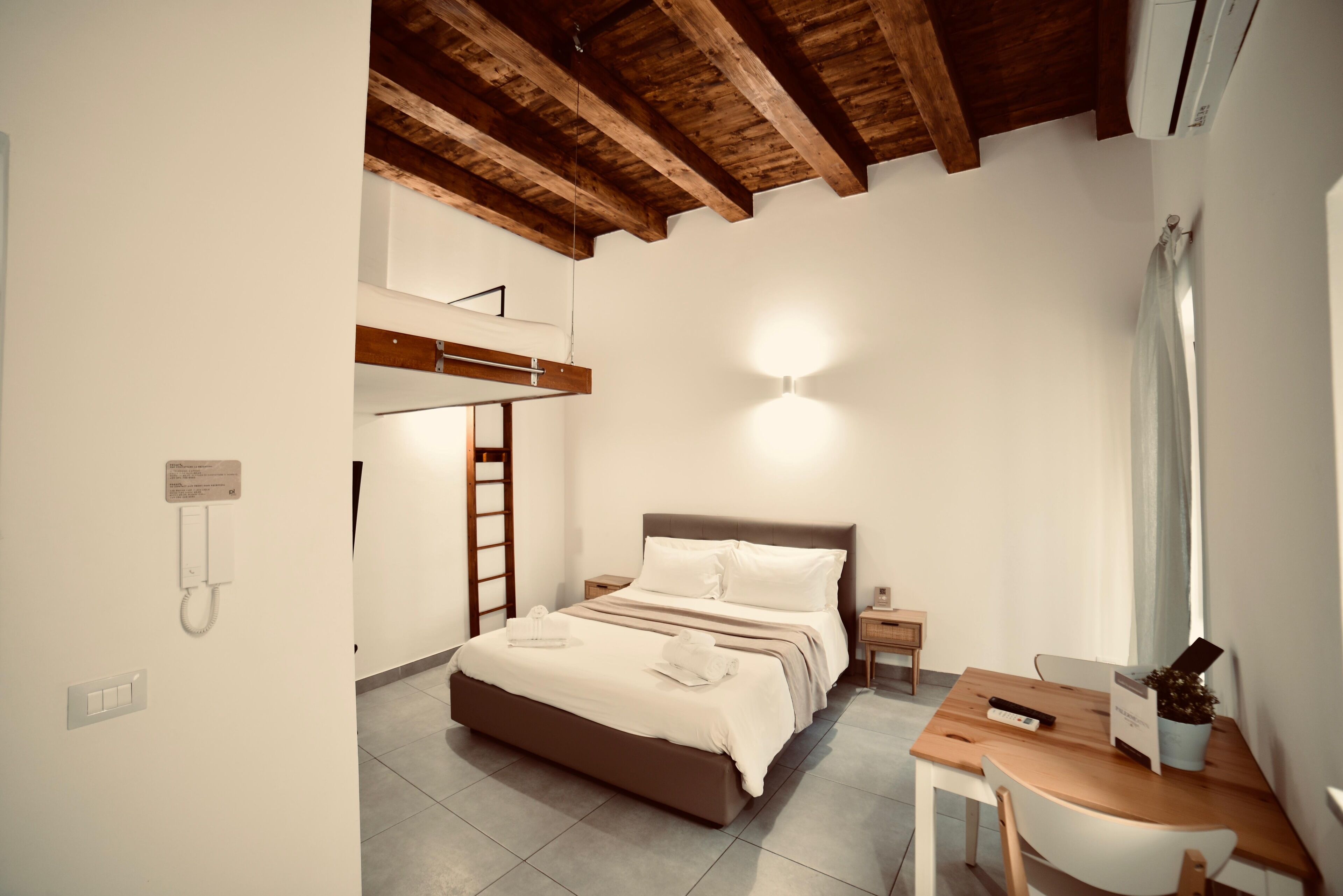 A bedroom with a bed, bedside table, and a desk with a chair. There is a ladder against the wall and a wall-mounted phone.