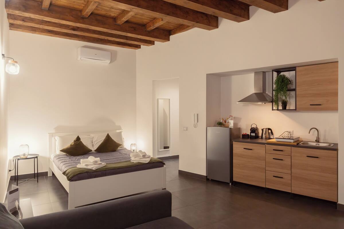 Un dormitorio moderno con una cama, una mesita de noche y una pequeña mesa auxiliar. Se observa una zona de cocina con un refrigerador, un lavabo y armarios.
