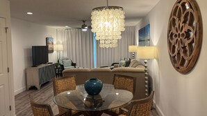 Un salon moderne avec une table à manger au plateau en verre, des chaises en osier, un canapé beige, un téléviseur et un élément décoratif en bois fixé au mur.