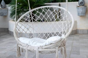 Un fauteuil suspendu en osier blanc, doté d’un coussin, placé sur un sol carrelé et entouré de plantes et de pots décoratifs.