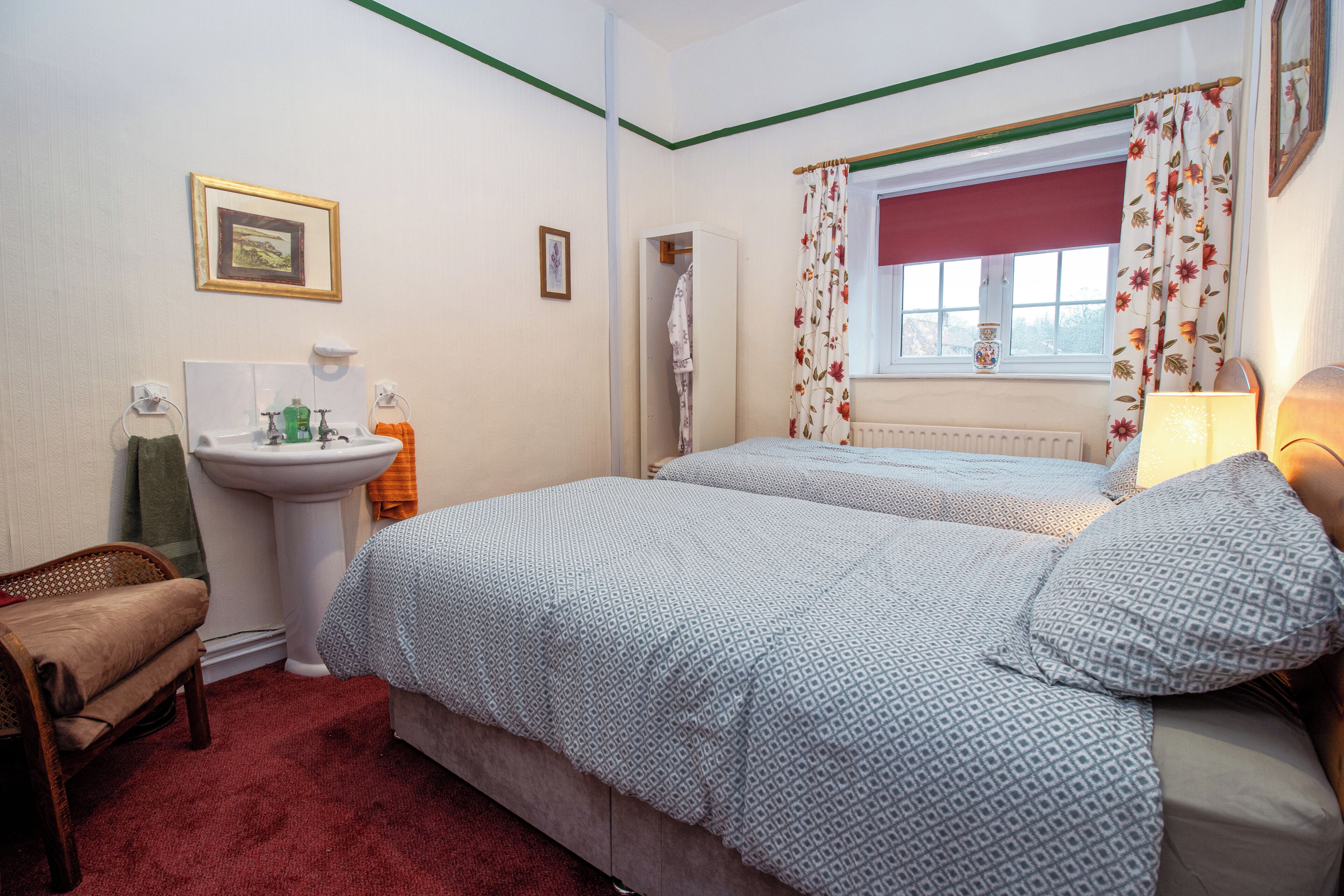 A bedroom with two beds, a bedside table, a chair, a window with curtains, a radiator, and a small picture on the wall.