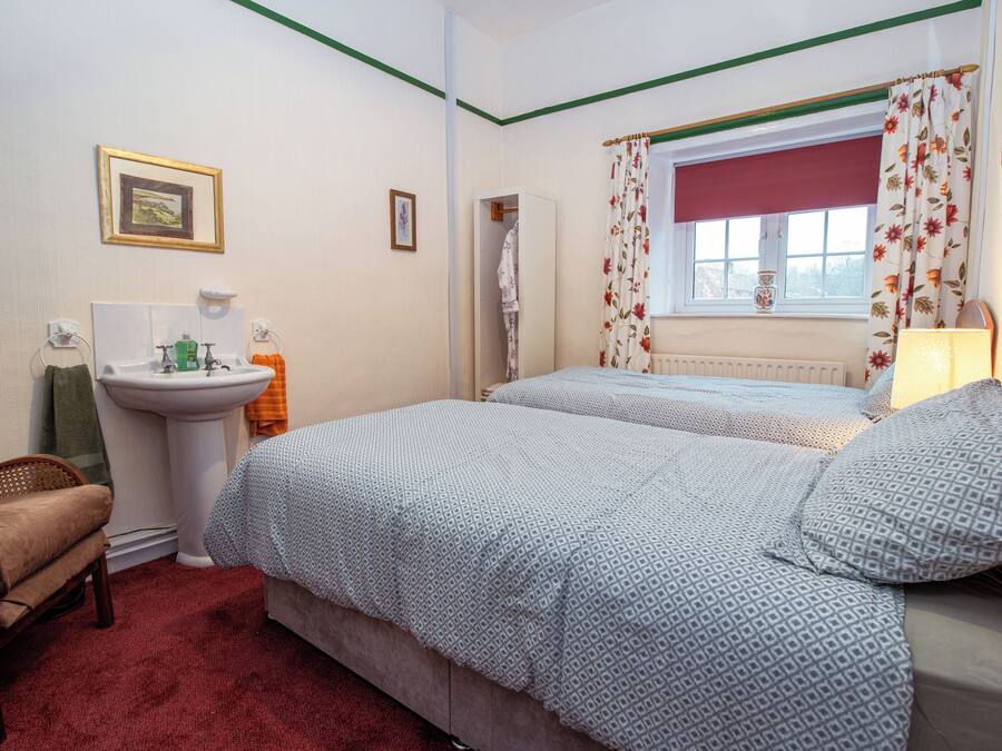 Une chambre avec deux lits, une table de chevet, une chaise, une fenêtre avec des rideaux, un radiateur et un petit tableau accroché au mur.