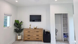 A modern living room with a ceiling fan, a flat-screen TV mounted on the wall, a wooden cabinet with drawers, a potted plant, and a black coffee machine.