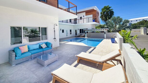 Ein moderner Poolbereich im Freien mit Liegestühlen und einer Sitzecke, umgeben von einem weißen Gebäude mit Holzdetails und großen Fenstern.