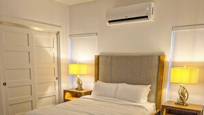 A bedroom with a wooden bed, two bedside tables with lamps, an air conditioning unit, and a door.