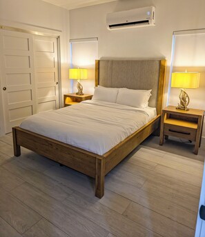 A bedroom with a wooden bed, two bedside tables with lamps, an air conditioning unit, and a door.