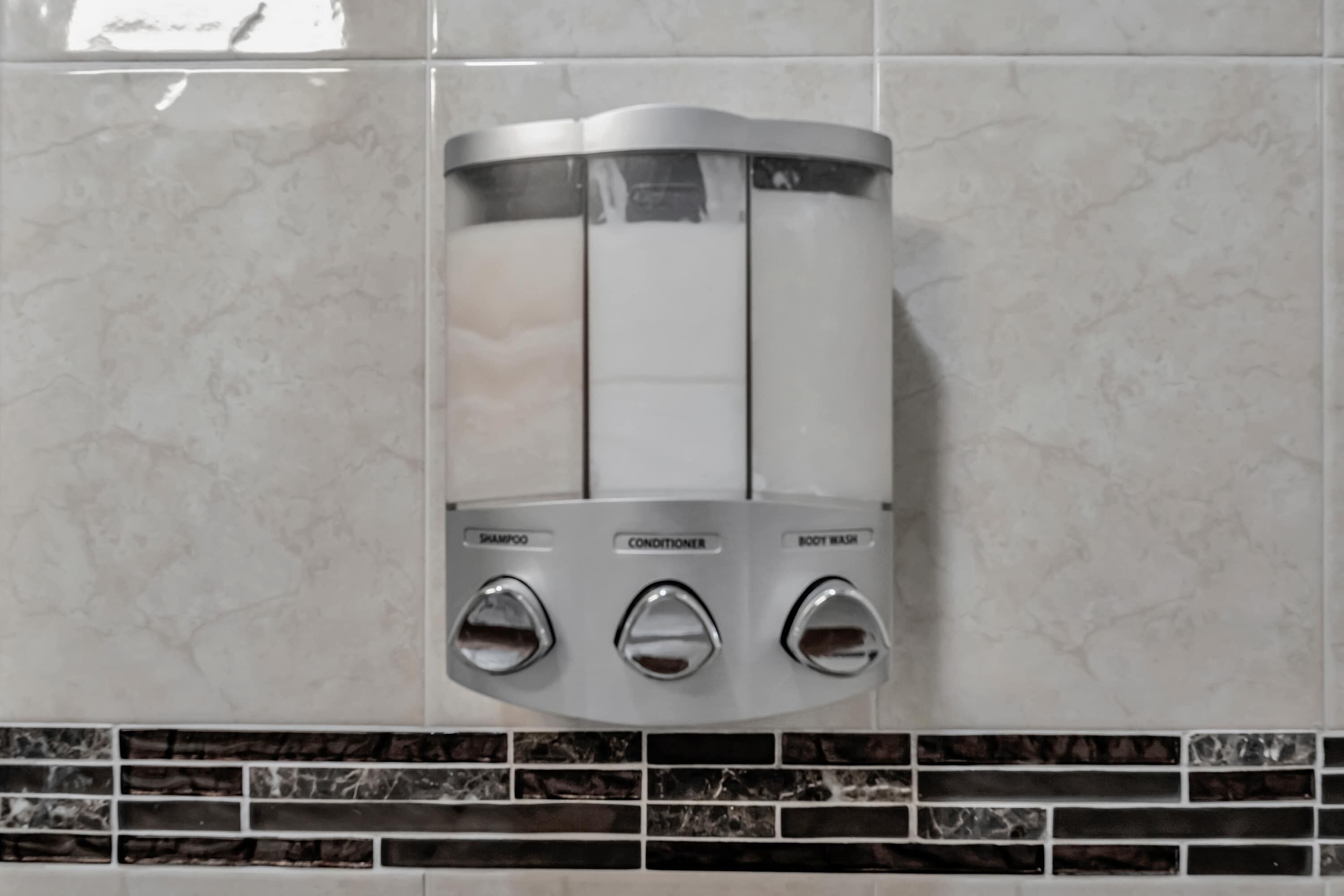 A wall-mounted dispenser with three compartments labeled for shampoo, conditioner, and body wash.