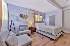 Ein Schlafzimmer mit einem Bett, zwei Stühlen, einem Nachttisch, einer Klimaanlage und einem an der Wand befestigten Fernseher.