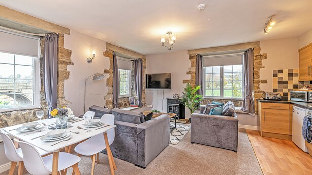 A modern living room with a dining area, stone walls, a flat-screen TV, and a kitchen with wooden cabinets. - Bridge End Mill Apartments by Guestz (Settle)