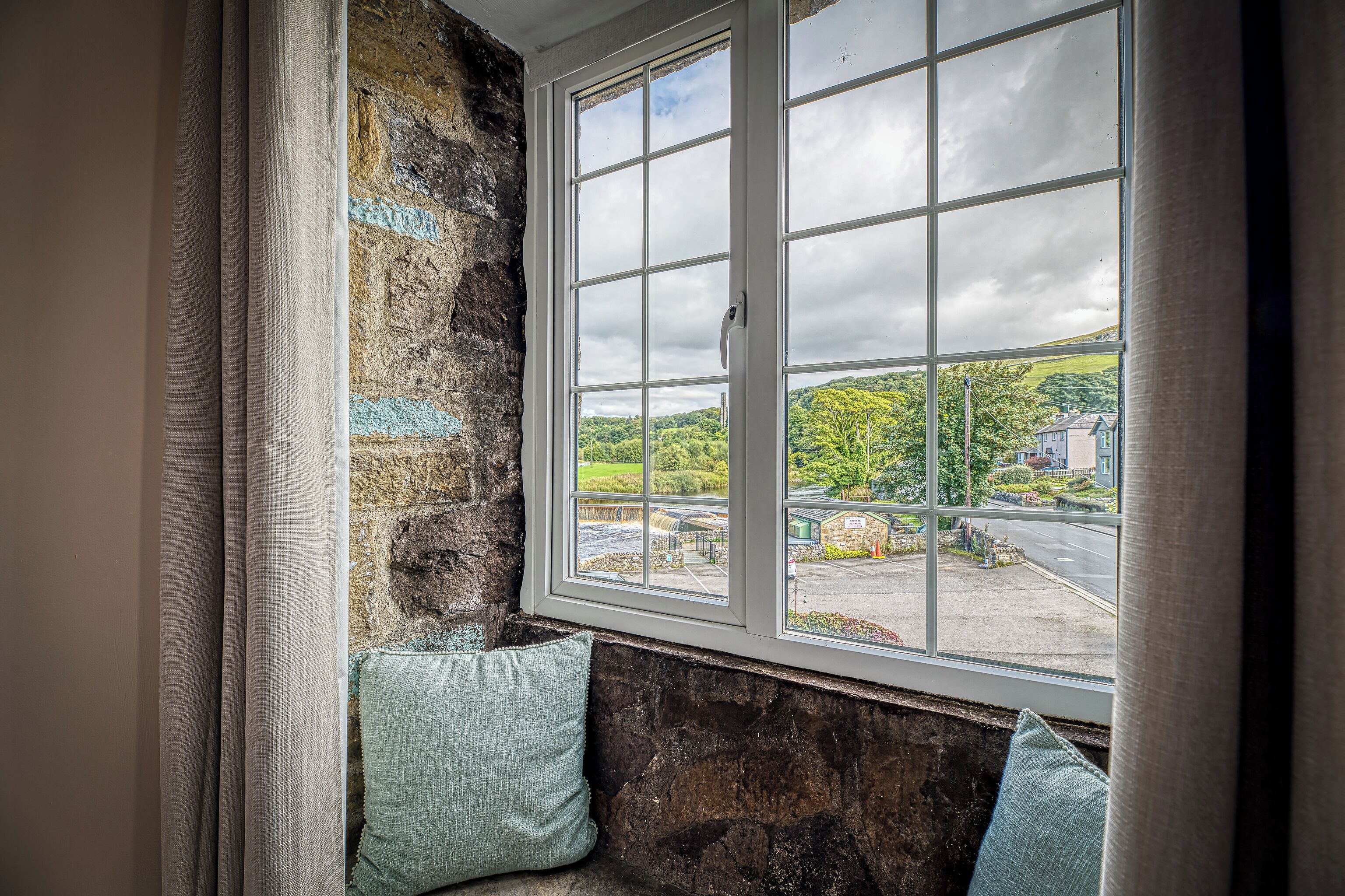 A room with a stone wall, a window with a view of a green landscape and buildings, and two pillows on a bench.