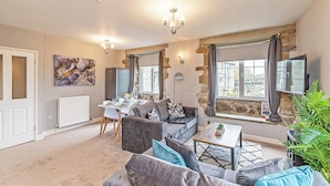 A modern living room with a sofa set, a dining table, and a large window with curtains. - Bridge End Mill Apartments by Guestz (Settle)