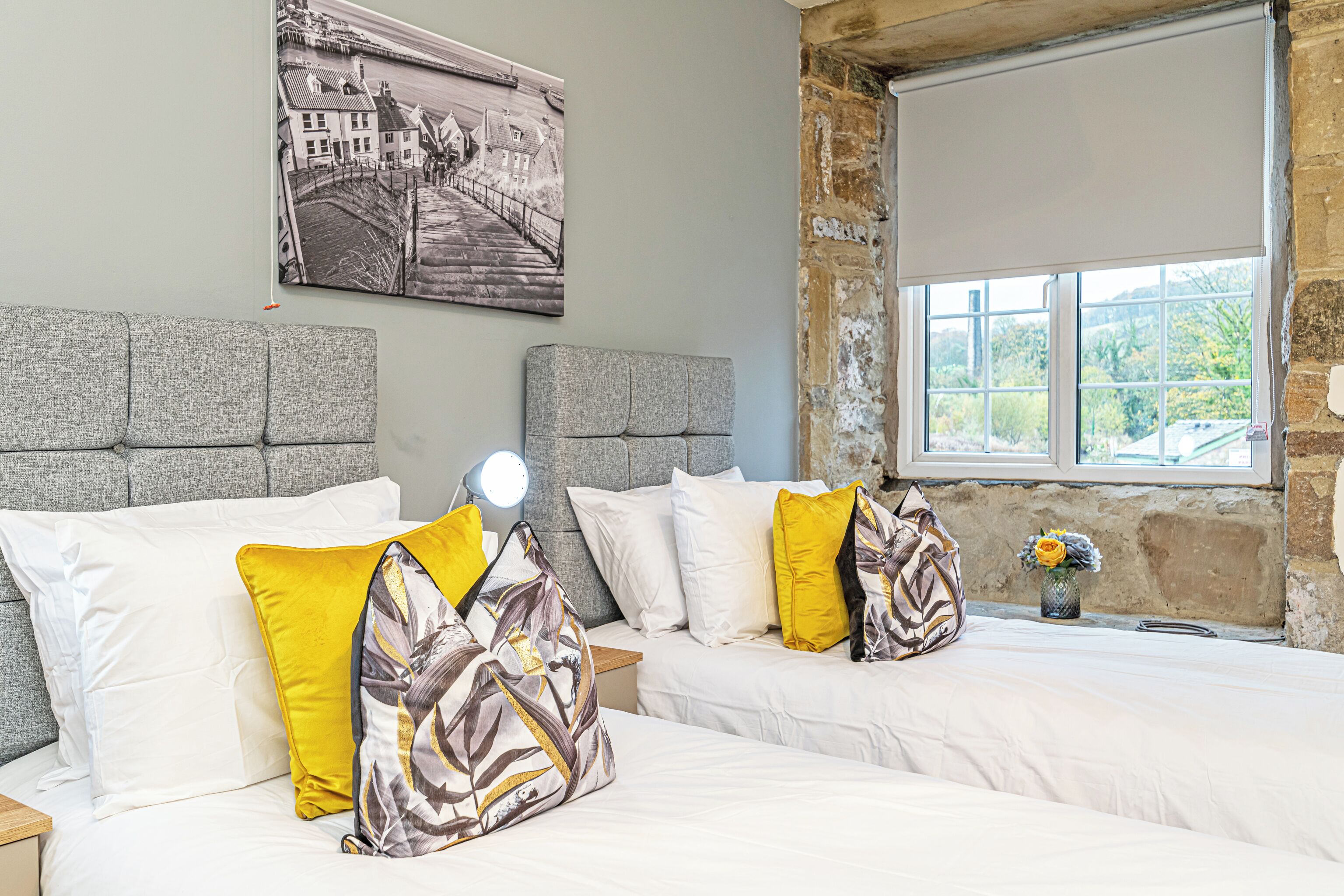 A hotel room with two beds, a large window with a view, a painting on the wall, and a nightstand with a lamp.